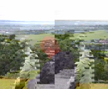 A male teacher standing in the countryside, writing on a clipboard. There is a lovely rural view behind him with green fields and trees.