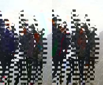 A group of young women standing on a rock in the outdoors with their arms reaching int the air, clearly enjoying geography