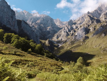 Mountain valley with steep rocky peaks, green slopes and scattered trees under a partly cloudy sky.