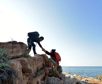 Two hikers on a seaside cliff on a sunny day. One is assisting the other by taking their hand and pulling them up. 