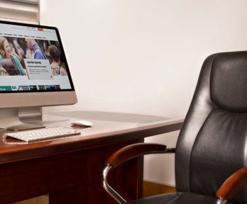 A leather office chair with wooden arm rests and a desk. A monitor on the desk is displaying the RGS website.
