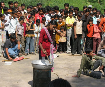 A large group of people gathered around a person with a metal bowl placed on a metal barrel. 