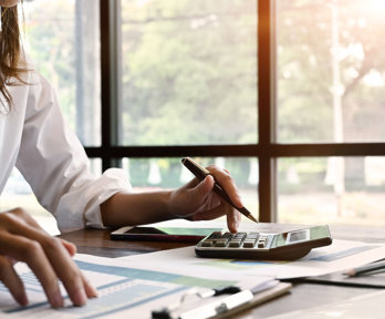 A person in a white shirt sat at a desk whilst typing on a calculator and looking at paper on a clip board.
