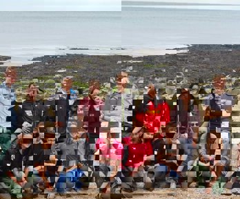 A group of older students lined up on a beach. In the background you can see a rocky beach with seaweed, and then the sea.