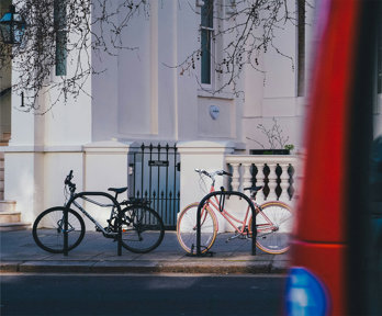 Two bicycles locked to racks outside a white townhouse, with a red double-decker bus passing in the foreground.