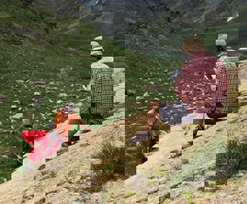 A researcher sitting on a rocky slope in a valley writing in a notebook while two others stand testing the rock hardness.