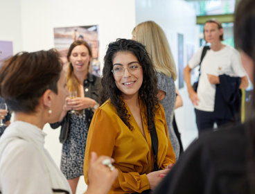 A person stands amongst a group of people chatting and drinking in a brightly lit exhibition space.