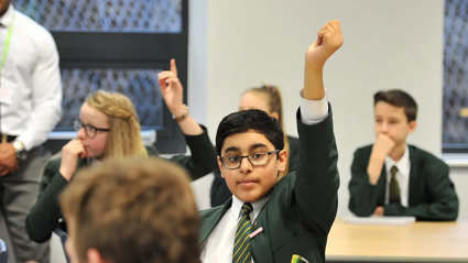 A young child wearing a school uniform sits in a classroom with their hand raised above their head to indicate they want to ask a question.