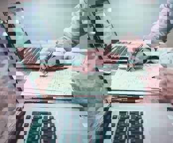 Two people sitting at a table writing on a piece of paper between laptops 