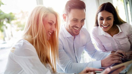 Three people sitting at a table looking at screens whilst smiling.
