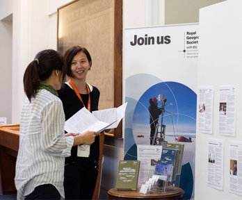 Two people chatting in front of a board of the Royal Geographical Society that says join us. 