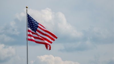 Flag of the USA on a pole waving against cloudy backdrop.