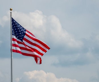 Flag of the USA on a pole waving against cloudy backdrop.