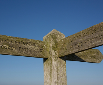 Wooden signpost showing three directions in English and Welsh. The left arrow says 'coast path' and the right arrow says 'public footpath'.